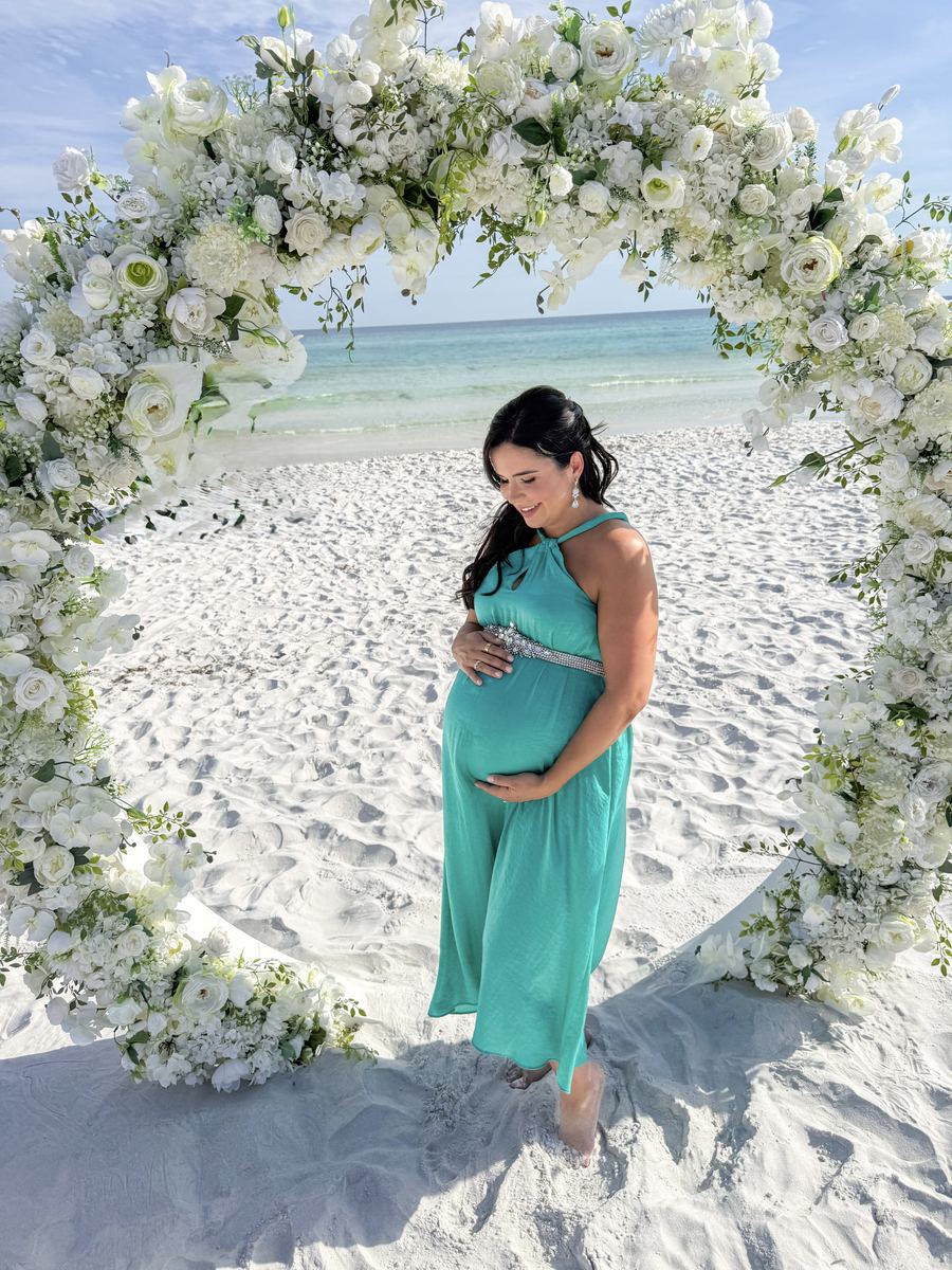 Rafaela pregnant in a teal dress under a white flower arch on the beach